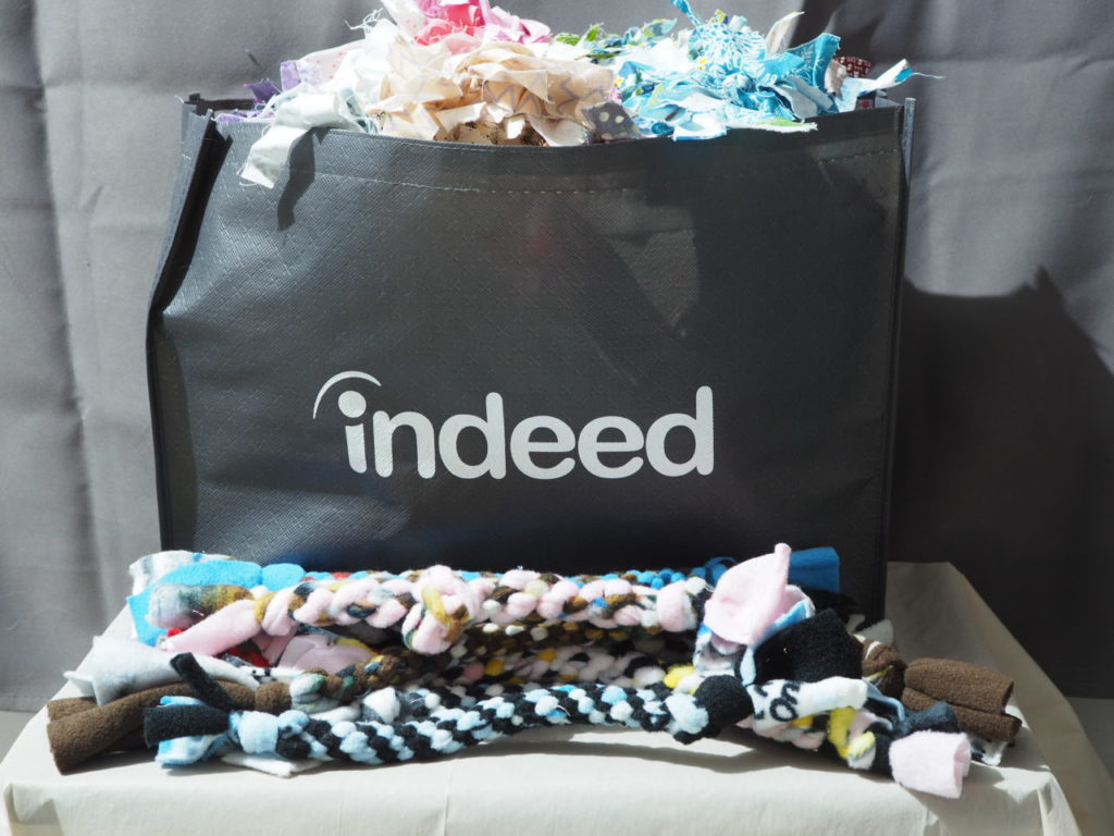 A tote bag filled with handmade fabric cat toys sits on table. In front of the bag there are handmade braided fleece dog toys. The tote bag has an logo on the front that says indeed.